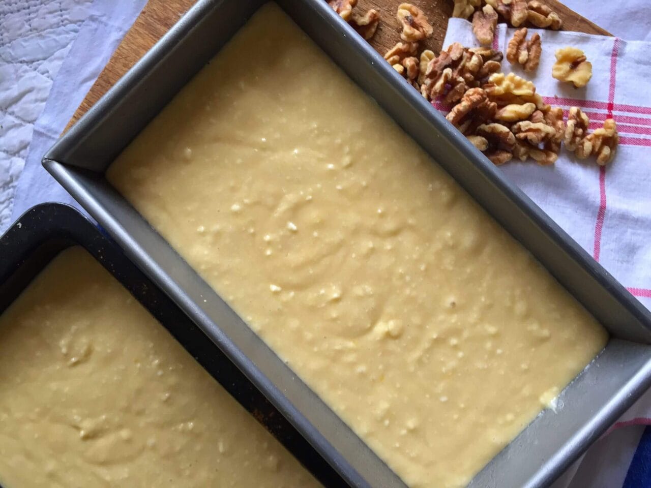 Two loaf pans filled with cake batter, next to a few scattered walnuts on a kitchen towel.