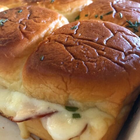 Close-up of slider buns topped with melted cheese and sprinkled with chopped herbs, with slices of ham visible inside, in a baking dish.
