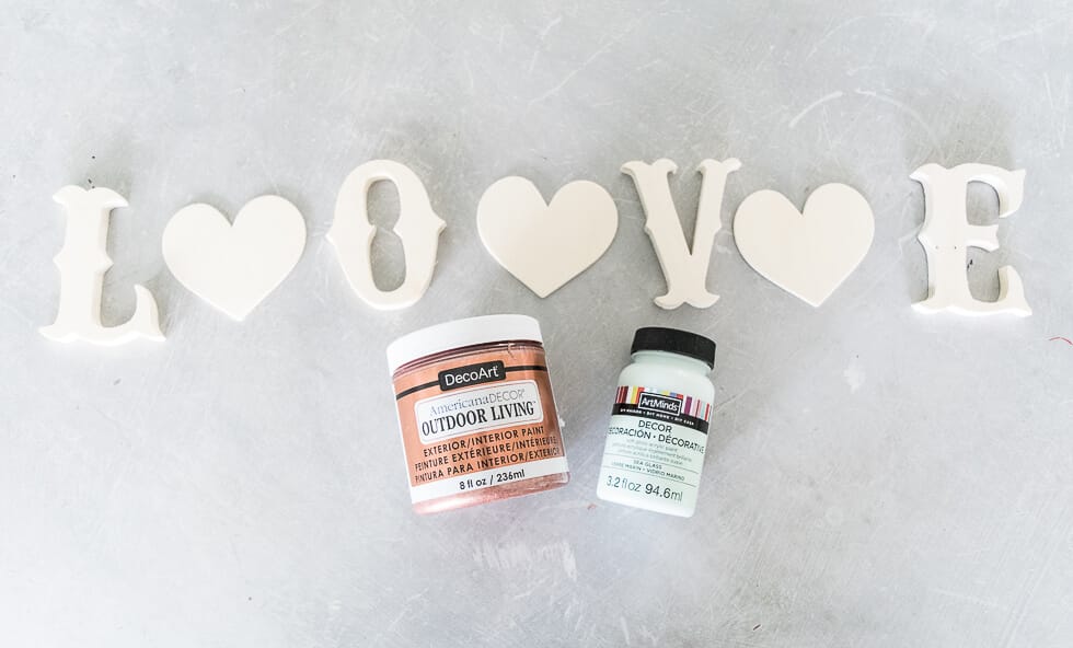 Wooden letters spelling "LOVE" with heart shapes in place of "O" and "V," alongside jars of DecoArt paint, arranged on a light gray surface.