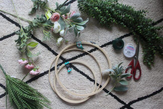 A flat lay of craft supplies on a textured rug, including embroidery hoops, artificial flowers, greenery, scissors, and floral wire.