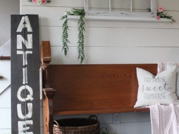 A wooden bench with a "FARMHOUSE sweet FARMHOUSE" pillow and a blanket sits next to a tall "ANTIQUES" sign, surrounded by rustic decor.