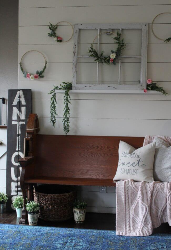 A rustic wooden bench with a "FARMHOUSE sweet FARMHOUSE" pillow and a blanket sits beneath a vintage window frame and decorative hoops with greenery, next to a tall "ANTIQUES" sign.