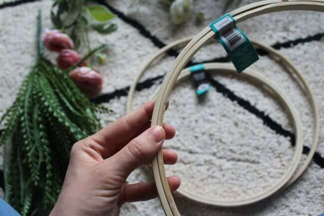 A hand holding an embroidery hoop, with artificial flowers and greenery laid out on a textured rug in the background.