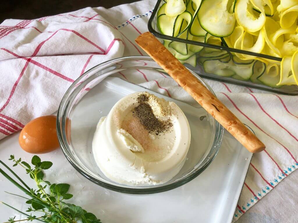 A bowl of ricotta cheese with spices on top, a wooden spoon, fresh herbs, an egg, and a dish of sliced zucchini and yellow squash on a cloth.