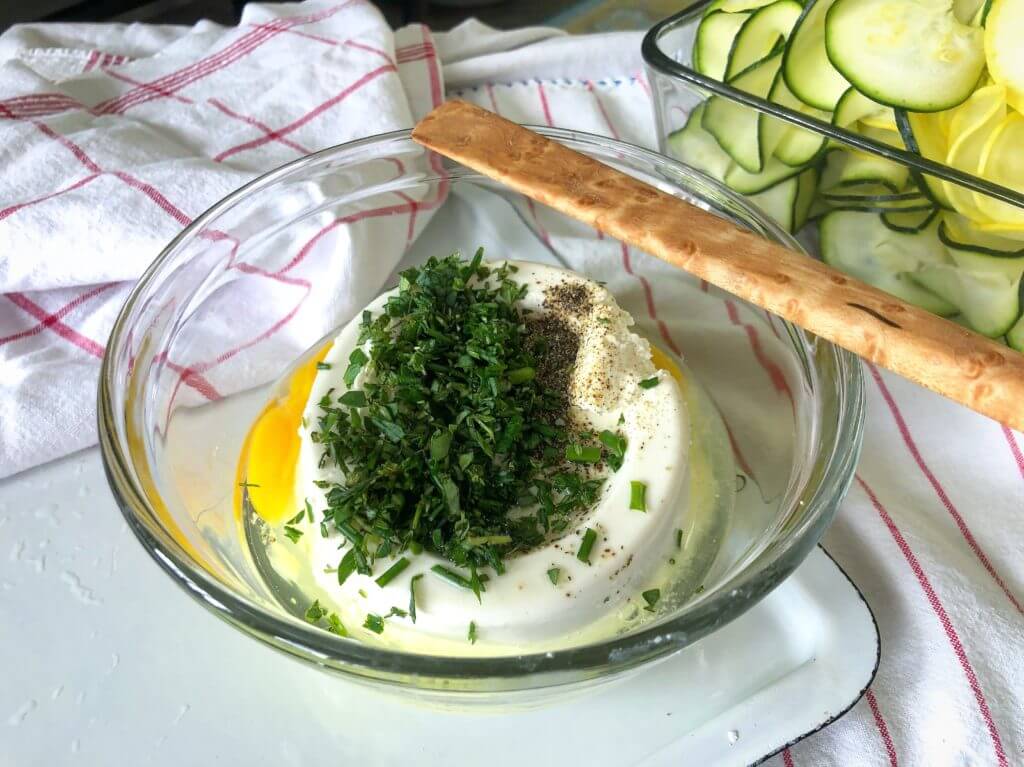 A bowl of ricotta cheese topped with fresh herbs and spices, a wooden spoon, and a dish of sliced zucchini and yellow squash on a cloth.