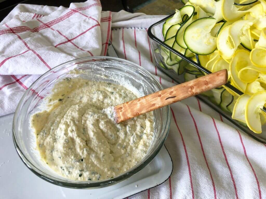 A bowl of creamy dip with a wooden spoon next to sliced zucchini and yellow squash on a cloth.