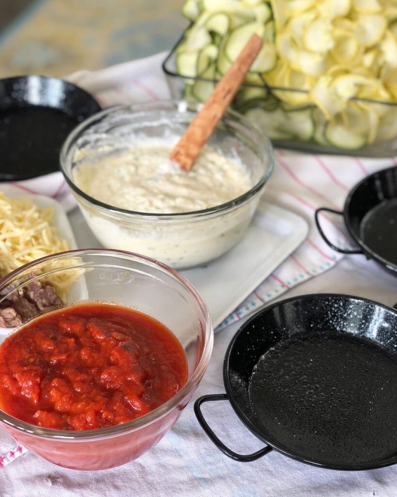 A bowl of tomato sauce, a bowl of creamy dip with a wooden spoon, shredded cheese, and sliced zucchini and yellow squash on a cloth, with two empty black pans.
