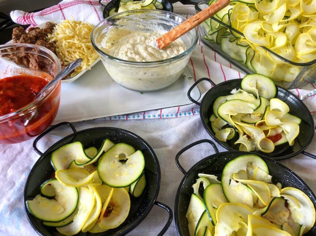 A variety of ingredients including tomato sauce, shredded cheese, seasoned ground meat, creamy dip, and sliced zucchini and yellow squash, with four small black pans partially filled with zucchini and squash.