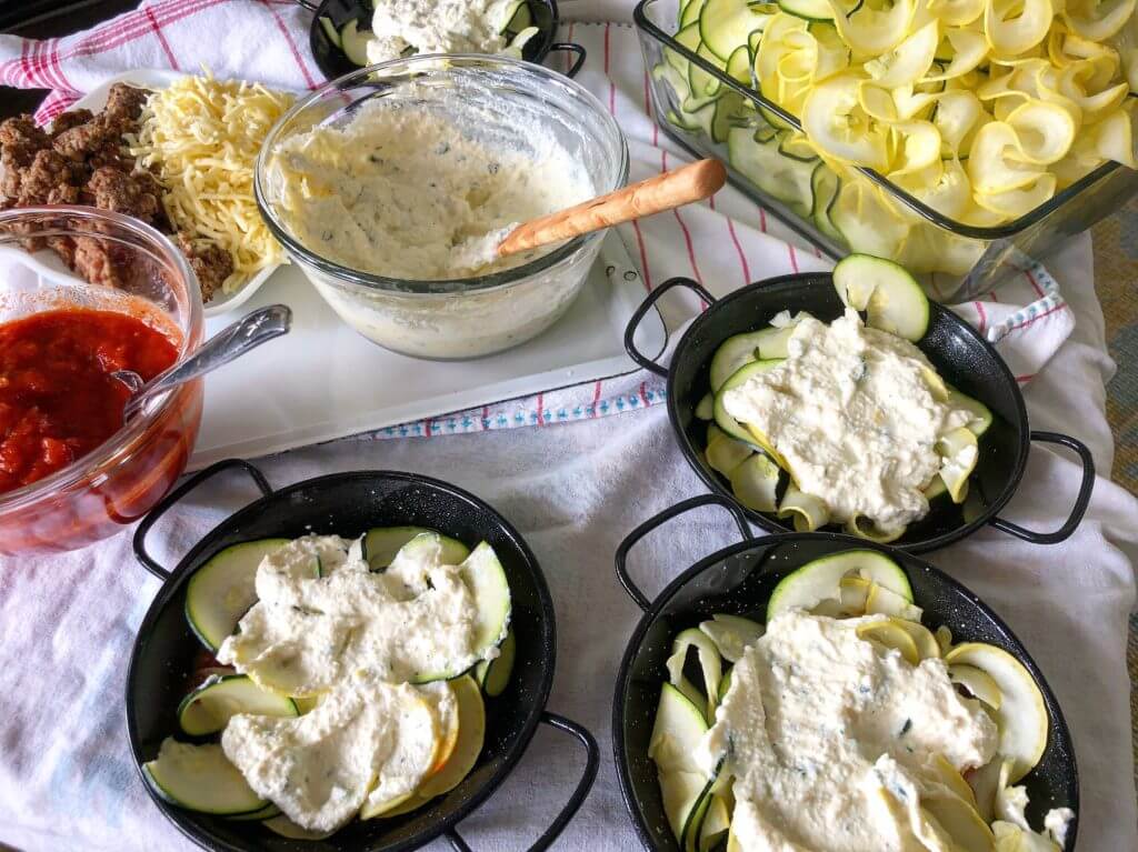 A variety of ingredients including tomato sauce, shredded cheese, seasoned ground meat, creamy dip, and sliced zucchini and yellow squash, with four small black pans partially filled with zucchini and squash.
