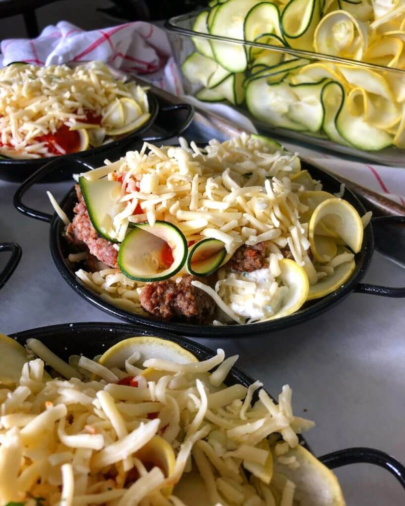 Small black pans filled with layers of zucchini, yellow squash, ground meat, tomato sauce, and shredded cheese.