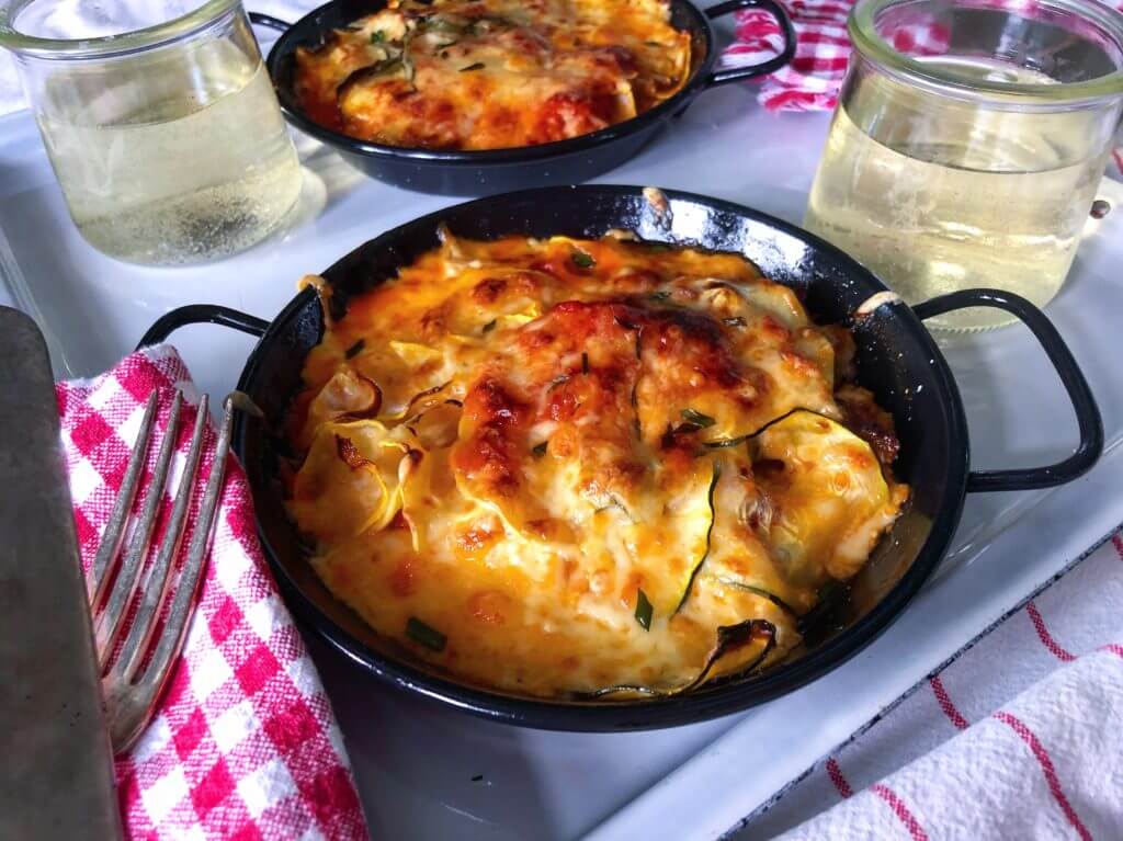 Baked zucchini and squash casserole topped with melted cheese in a small black pan, served with a glass of white wine.