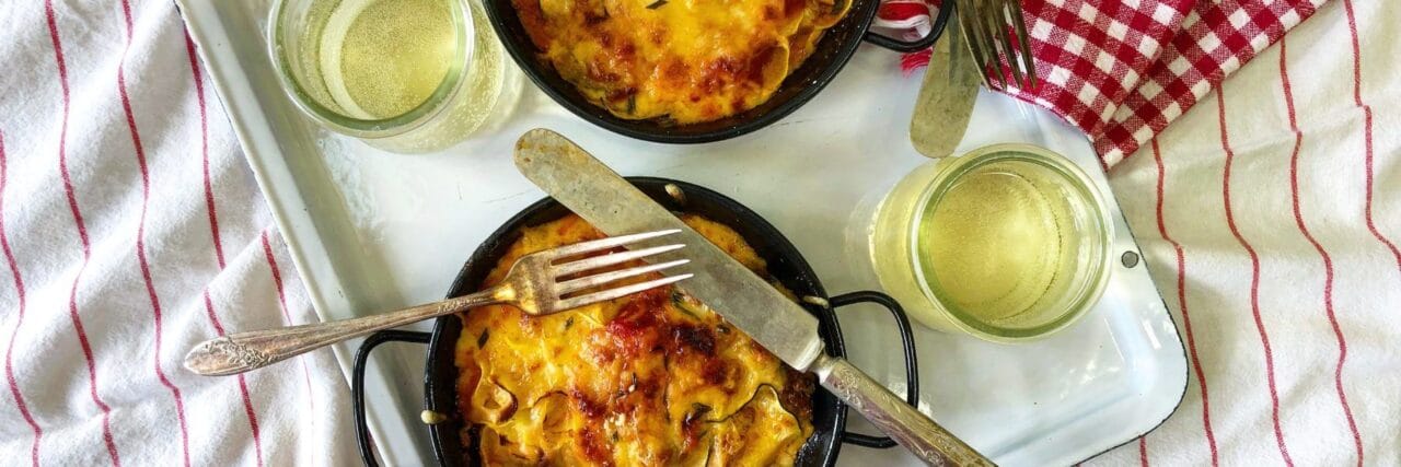 Two small black pans of baked zucchini and squash casserole topped with melted cheese, served with glasses of white wine on a tray with forks and knives.