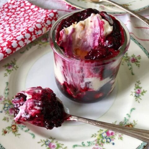 A glass jar filled with berry banana pudding, featuring layers of whipped cream and berry sauce, served on a decorative plate with a spoonful of the dessert beside it.