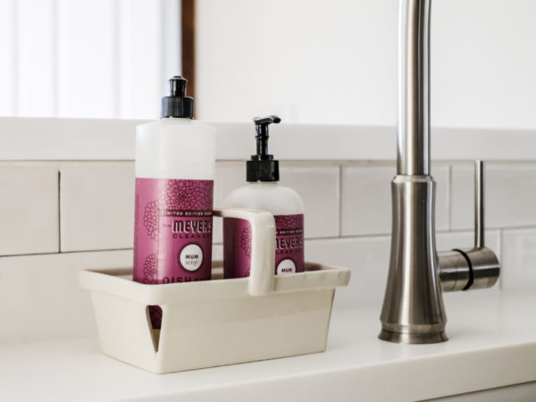 two bottles of meyers kitchen soap in a tray next to a white farmhouse sink with a stainless steel faucet