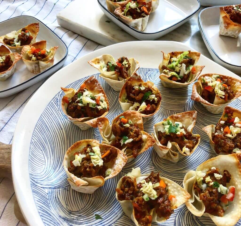 A platter of crispy wonton cups filled with a ground meat and vegetable mixture, garnished with chopped cilantro and red bell peppers, placed on a striped cloth background with additional filled wonton cups on smaller plates.