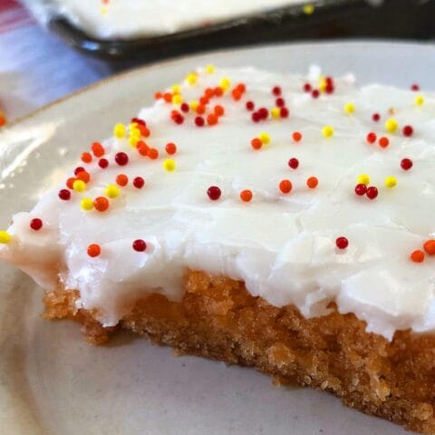 A slice of candy corn sheet cake topped with white frosting and colorful sprinkles on a white plate. The cake has a golden brown crust and moist, orange-colored interior.