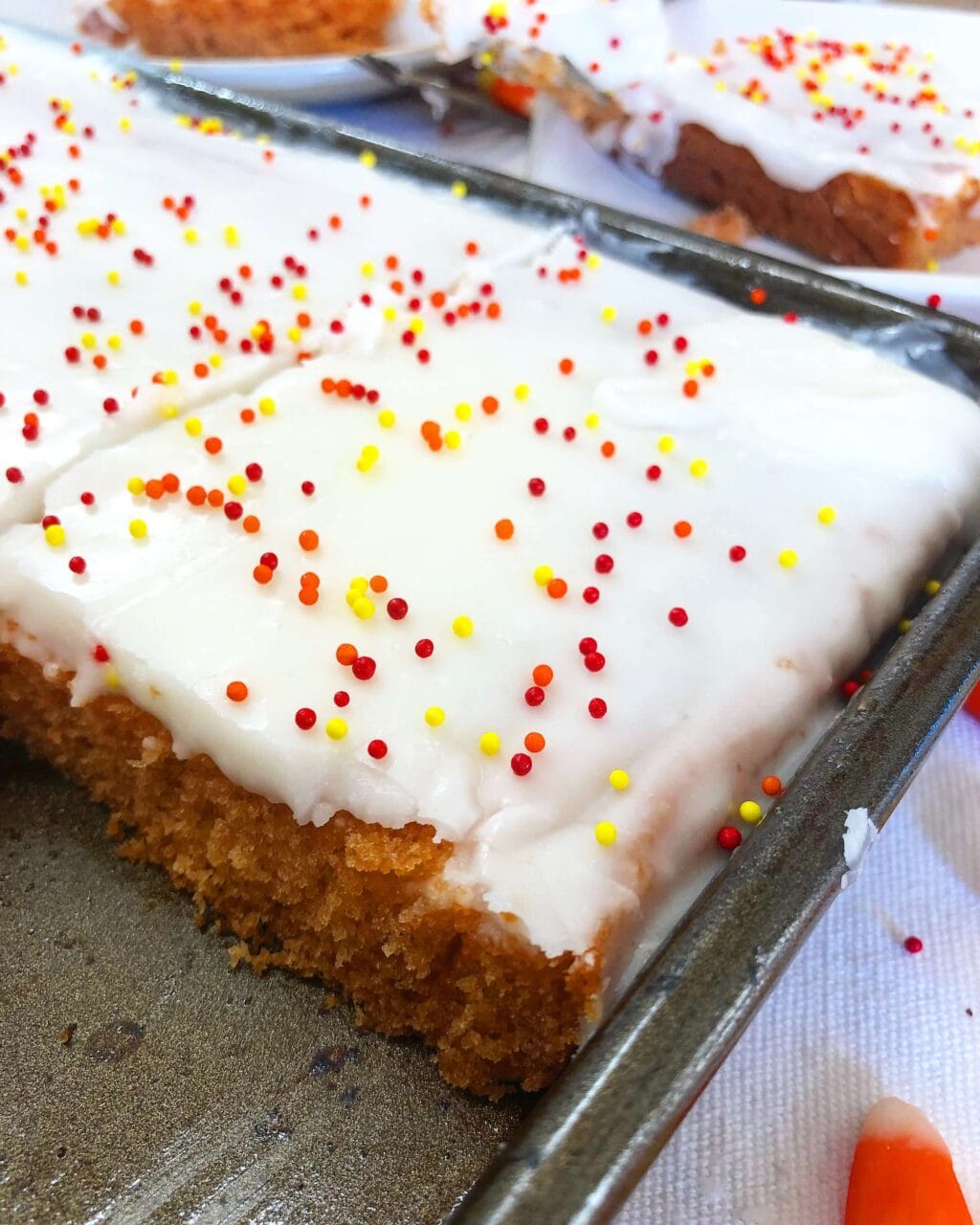 A close-up of a candy corn sheet cake with white frosting, topped with red, yellow, and orange sprinkles, cut into squares and displayed on a baking tray.