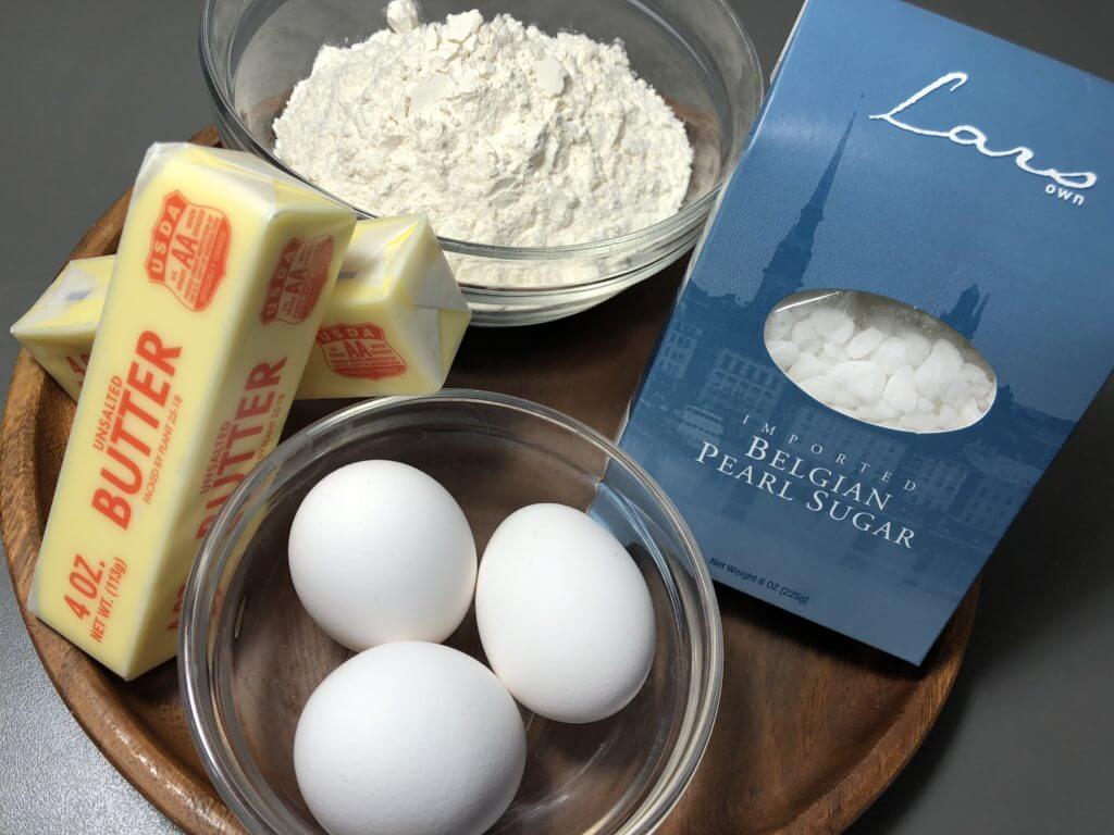 A wooden tray holding sticks of butter, a bowl of flour, a box of Belgian pearl sugar, and a bowl with three eggs.