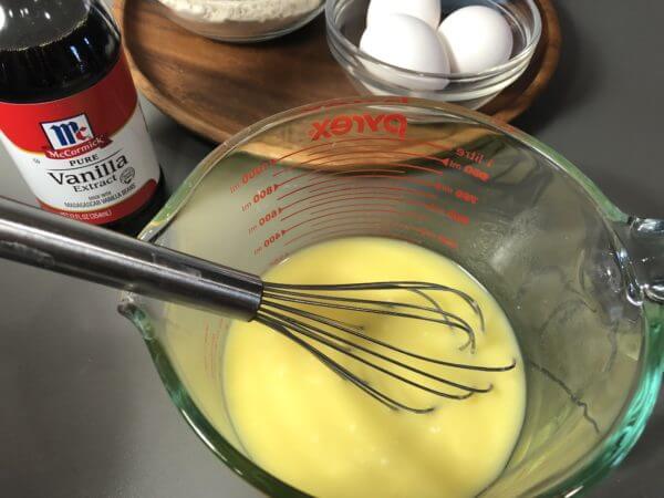 A Pyrex measuring cup with a whisk and a yellow liquid mixture next to a bottle of vanilla extract, a bowl of flour, and a bowl of eggs.