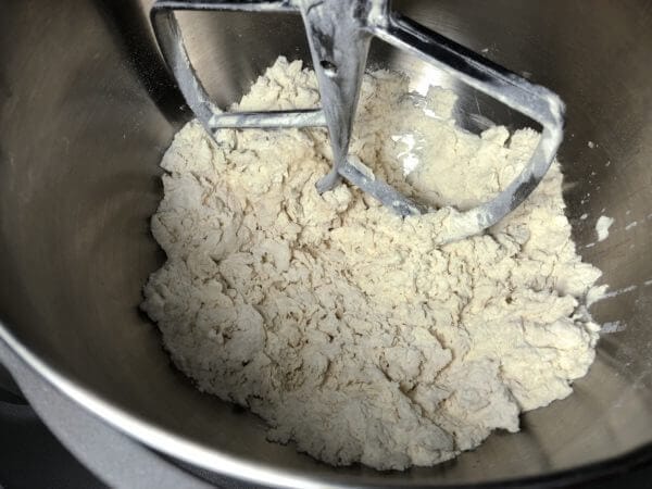 A stand mixer bowl with a paddle attachment mixing a crumbly dough.
