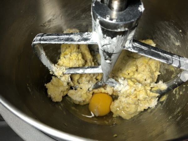 A stand mixer bowl with a paddle attachment mixing dough and a cracked egg.