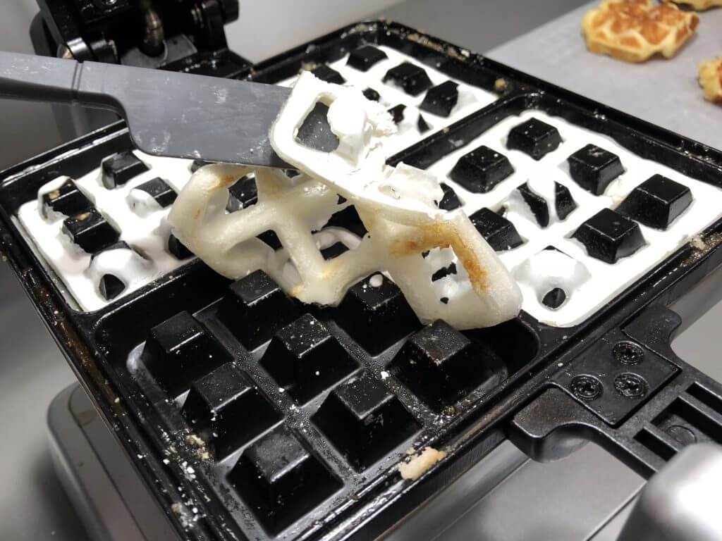 A spatula lifting a partially cooked waffle with batter stuck to the waffle iron.