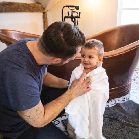 father drying off son with towel in front of a copper bath tub