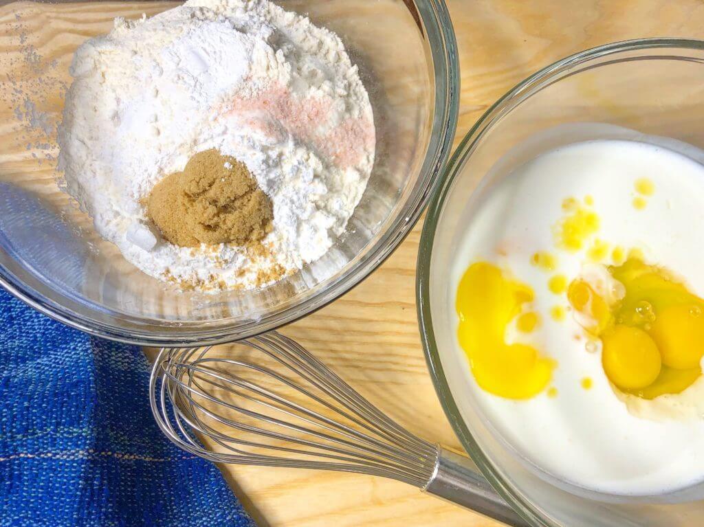 Two bowls with baking ingredients, one with flour and brown sugar, the other with eggs and milk, next to a whisk and a blue towel.
