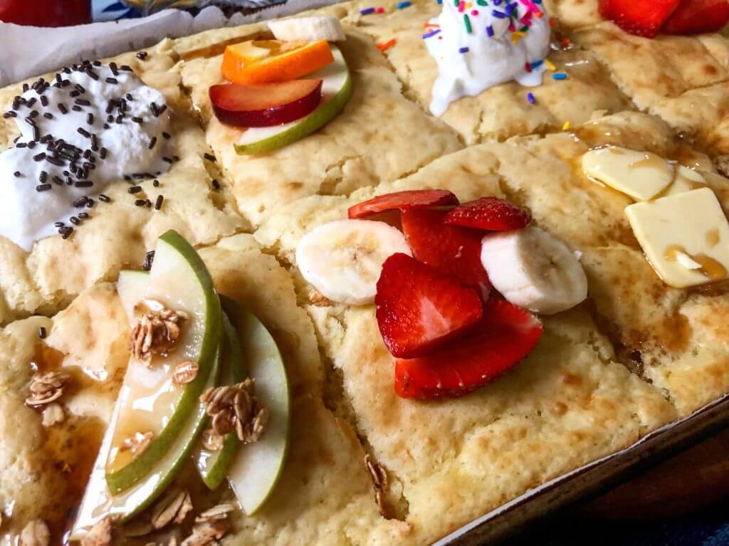Baked pancake squares topped with various ingredients, including whipped cream and chocolate sprinkles, sliced fruits, granola, and syrup.