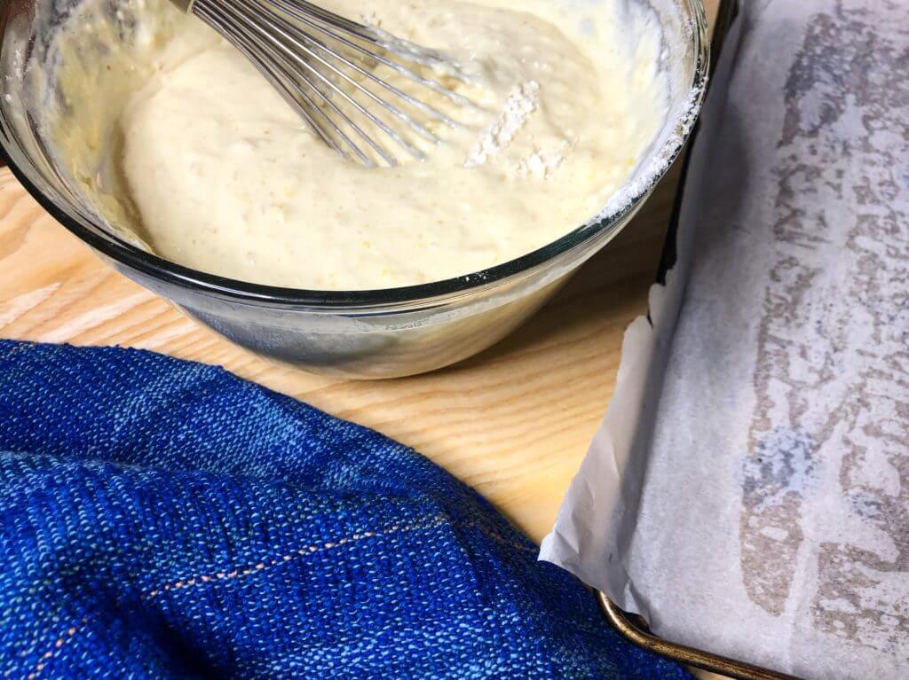 Bowl of batter with a whisk next to a lined baking tray and a blue towel.