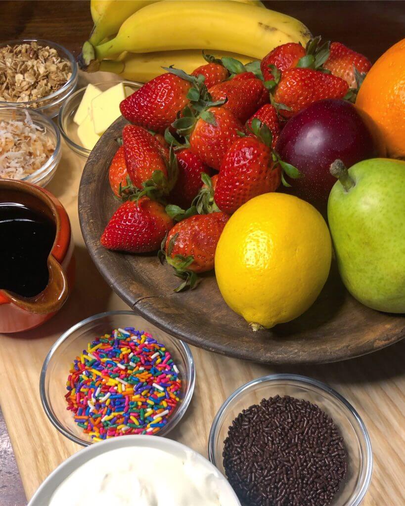 Assorted fruits including strawberries, bananas, lemons, and an apple, surrounded by small bowls of toppings like sprinkles, granola, and syrup.