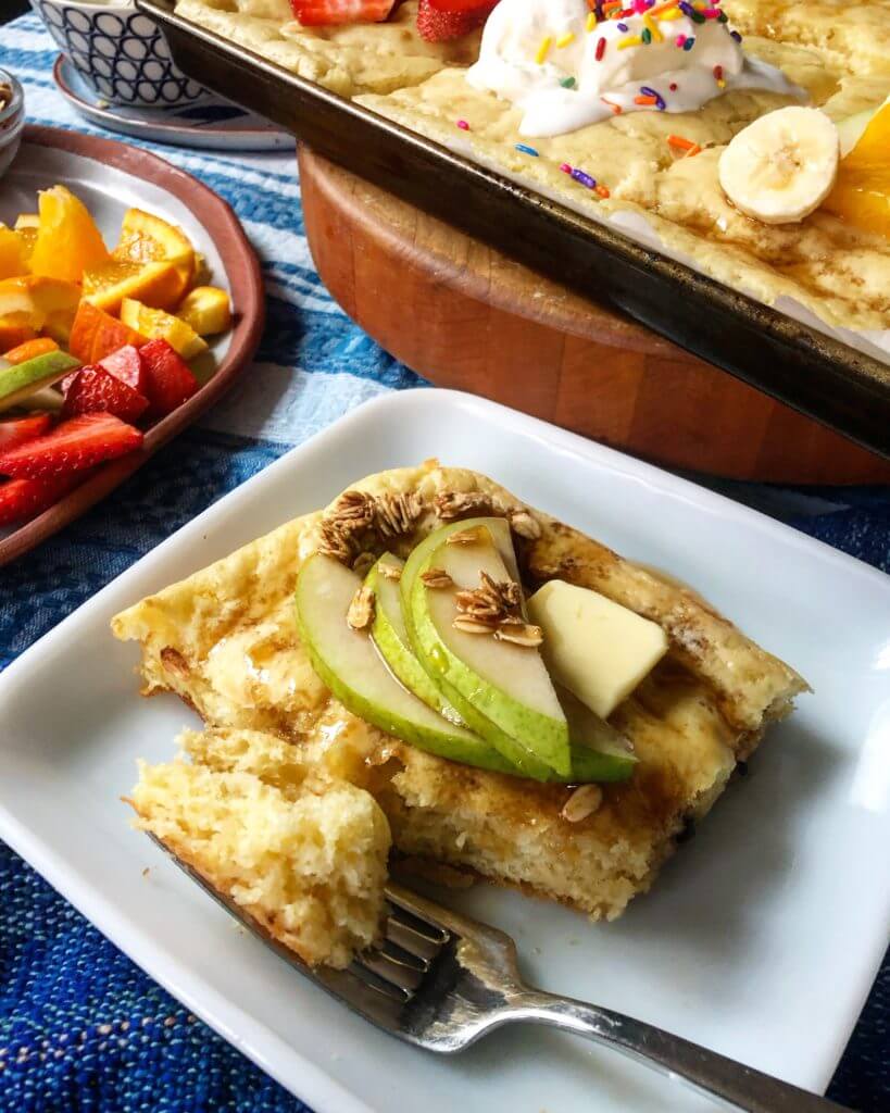 Slice of baked pancake on a plate topped with butter, syrup, and sliced green apples, next to a baking tray and a plate of mixed fruits.