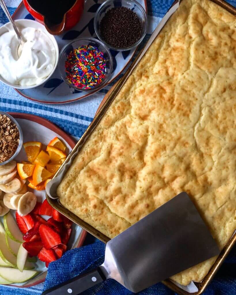Baked pancake in a tray with a spatula, surrounded by bowls of whipped cream, syrup, sprinkles, and a plate of sliced fruits.