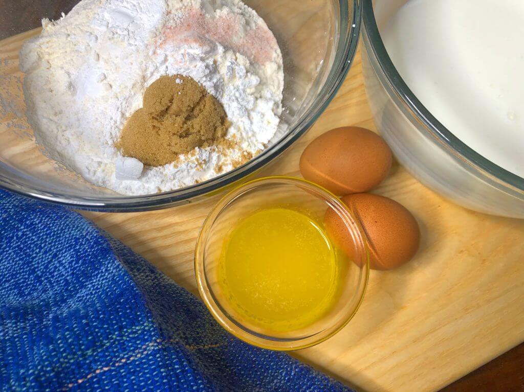 Ingredients for baking including flour, brown sugar, melted butter, eggs, and milk, next to a blue towel.