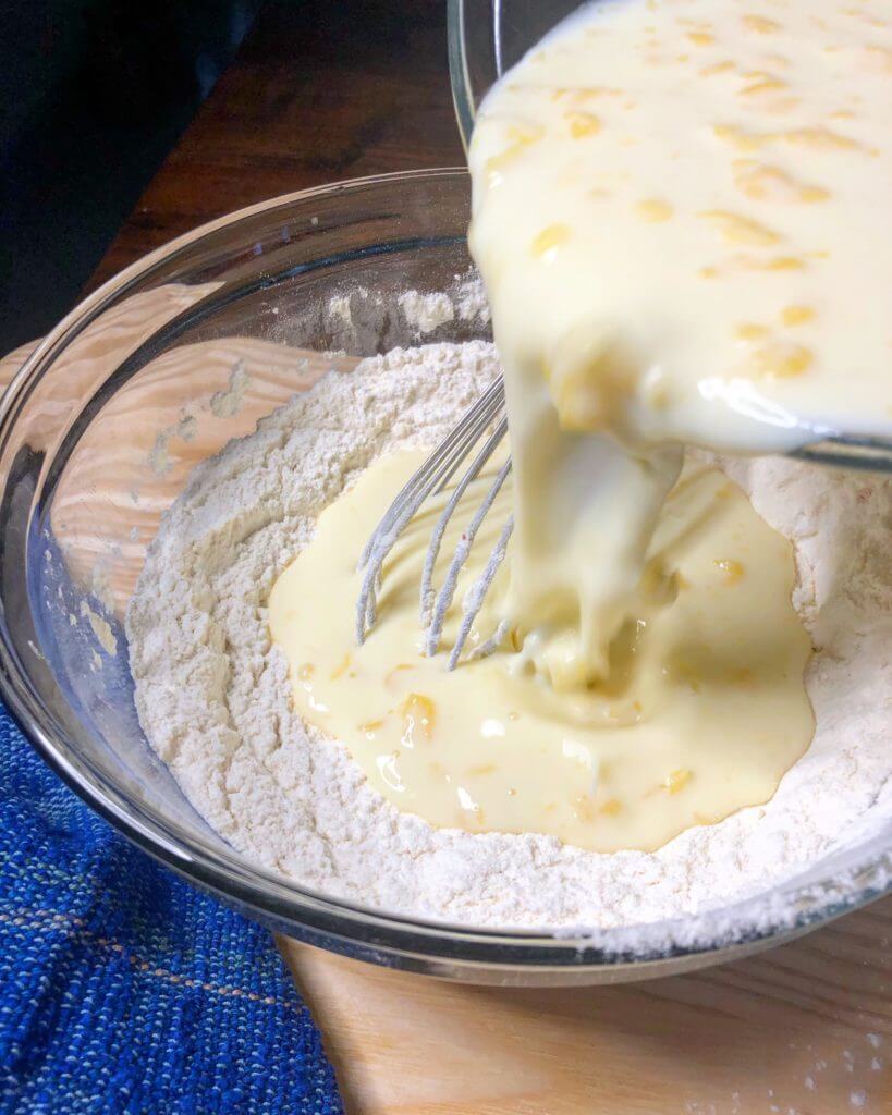 Mixing wet ingredients into dry ingredients in a bowl with a whisk, next to a blue towel.