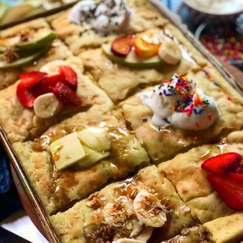 Baked pancake squares topped with various toppings including strawberries, bananas, whipped cream, sprinkles, and granola.