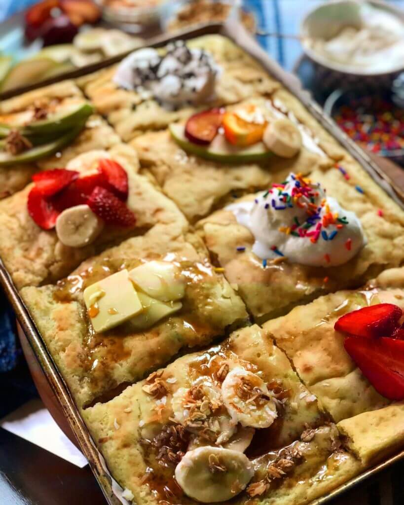 Baked pancake squares topped with various toppings including strawberries, bananas, whipped cream, sprinkles, and granola.