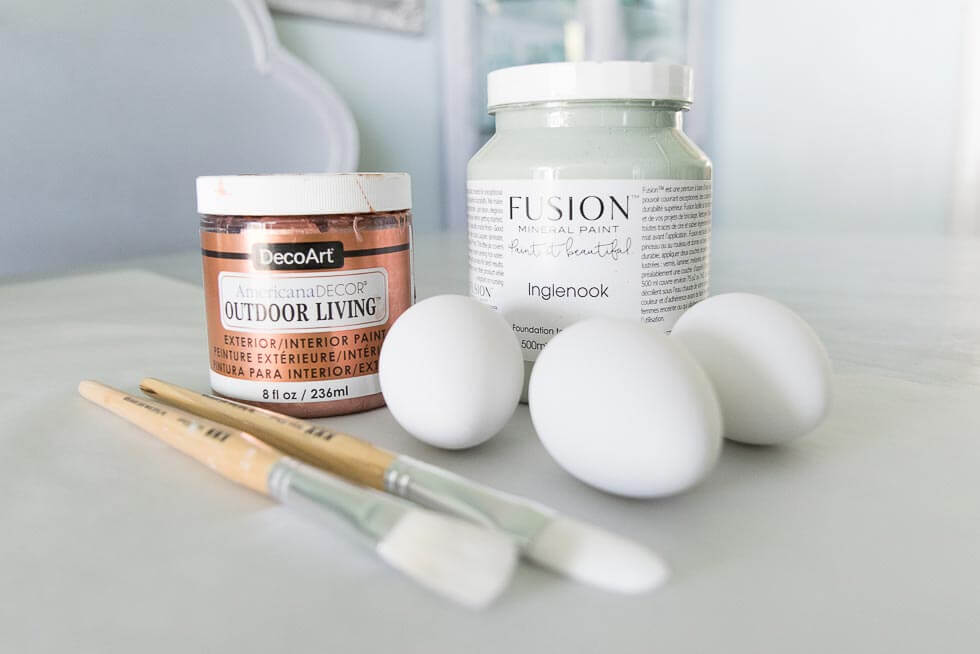 Two jars of paint, brushes, and three white eggs are arranged on a table, ready for a crafting project.