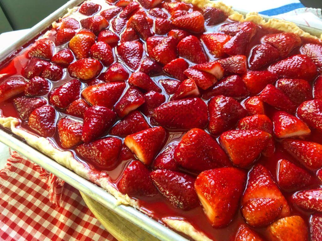 A close-up image of a strawberry pie with glistening fresh strawberries on top of a glossy, red filling in a rectangular pan.