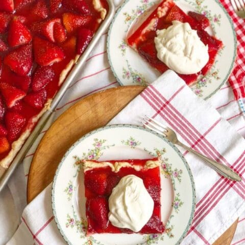A rectangular strawberry pie in a baking pan, with two plates each holding a slice of the pie topped with whipped cream.