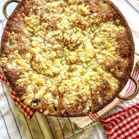 A golden-brown berry crumble cake in a baking dish sits on a red and white checkered cloth.
