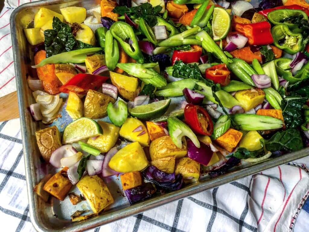 A baking sheet filled with a colorful assortment of chopped vegetables, including bell peppers, red onions, potatoes, lime wedges, and leafy greens, ready for roasting.