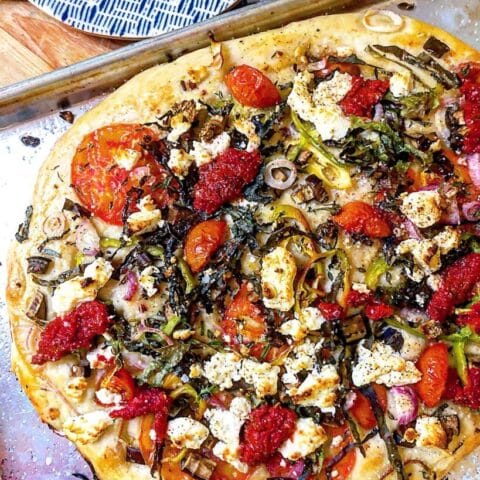 A freshly baked vegetable pizza with a golden crust, topped with tomatoes, onions, greens, dollops of tomato sauce, and crumbled cheese on a baking sheet.