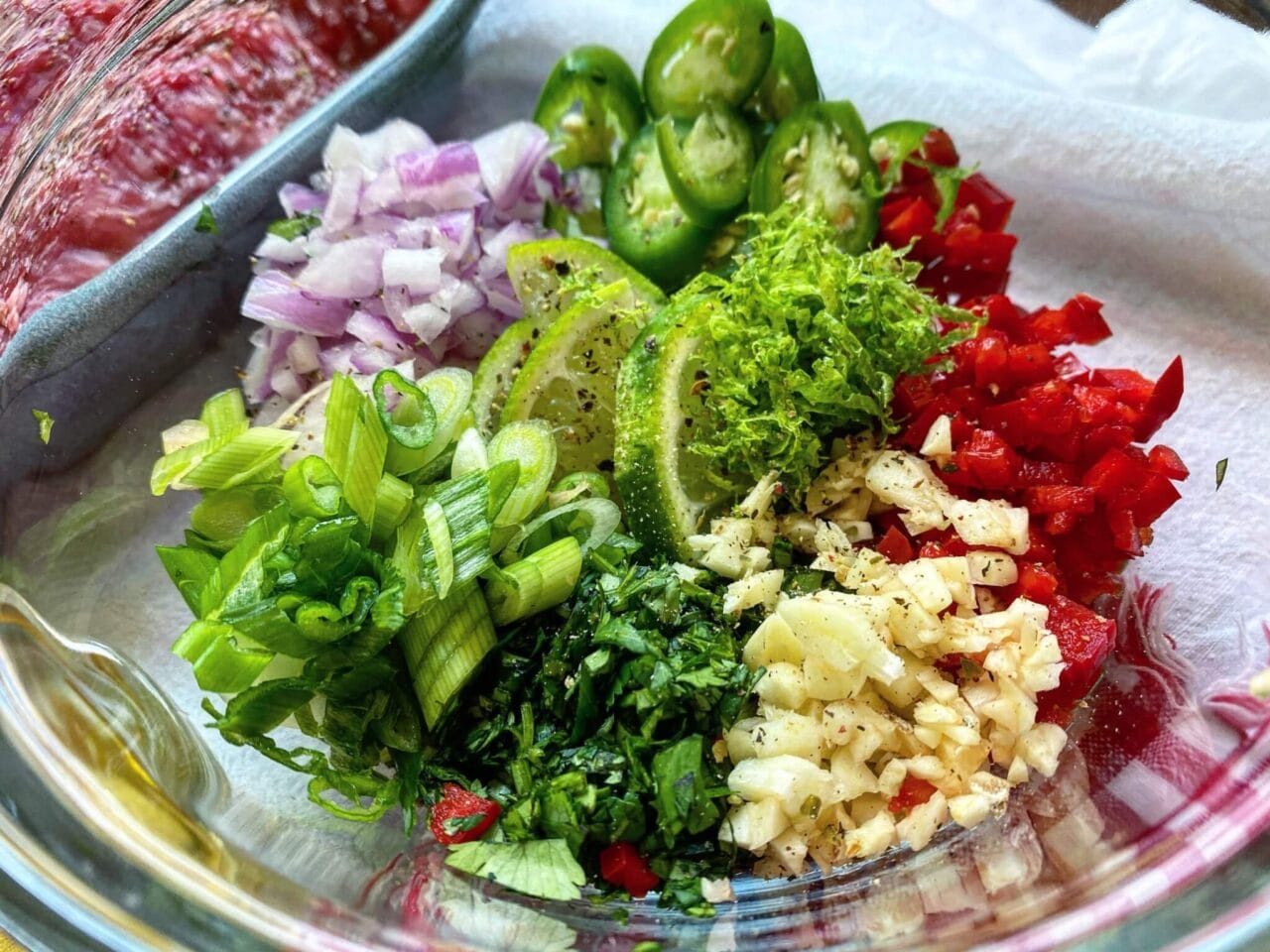 Chopped vegetables including red onions, green onions, jalapeños, red peppers, garlic, cilantro, and lime zest in a glass bowl.