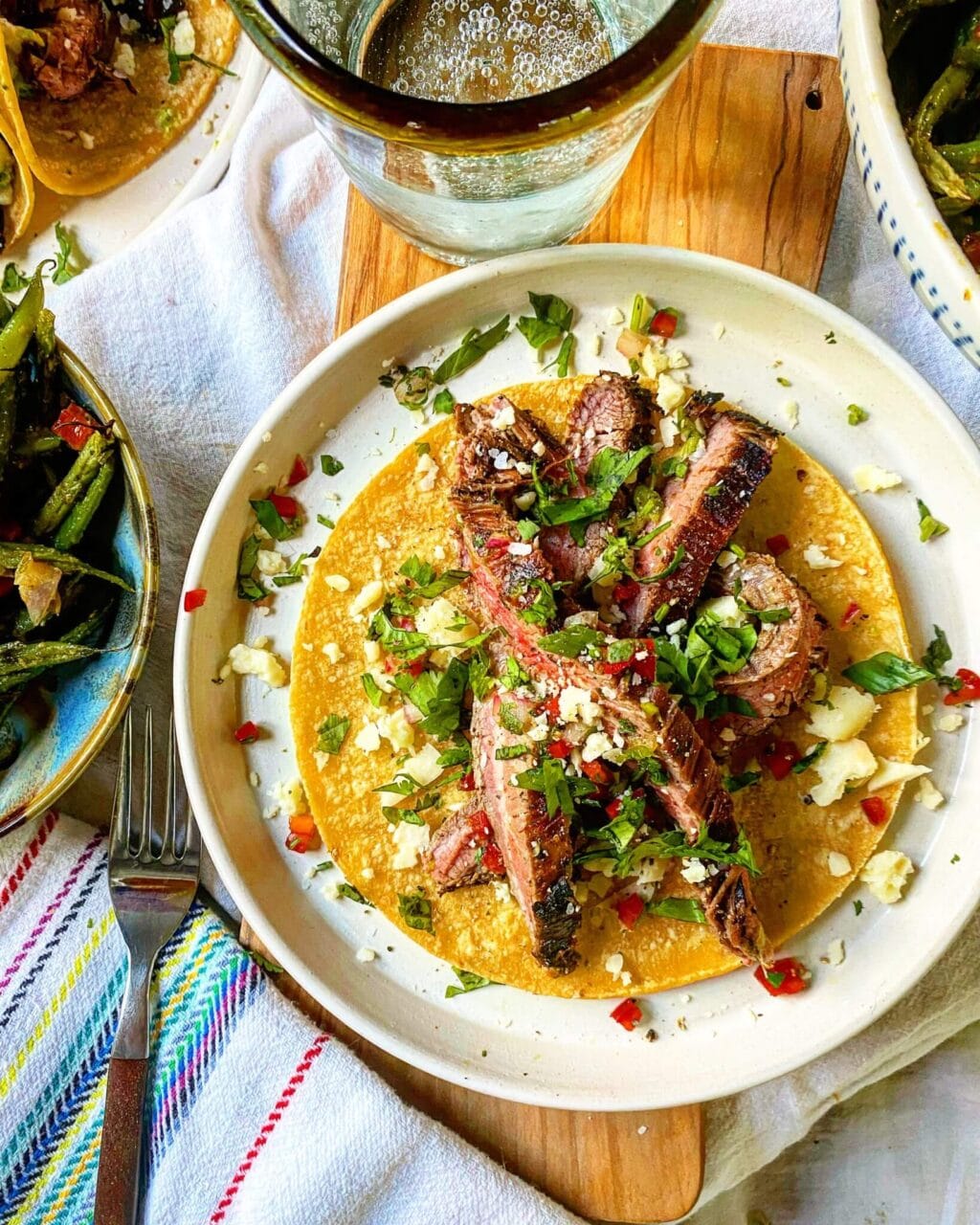 A plate with a taco filled with grilled meat strips, cilantro, red peppers, and crumbled cheese, accompanied by a glass of sparkling water.