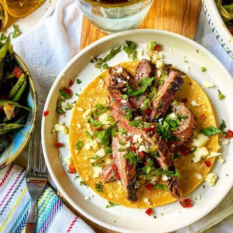 A plate with a taco filled with grilled meat strips, cilantro, red peppers, and crumbled cheese, accompanied by a glass of sparkling water.