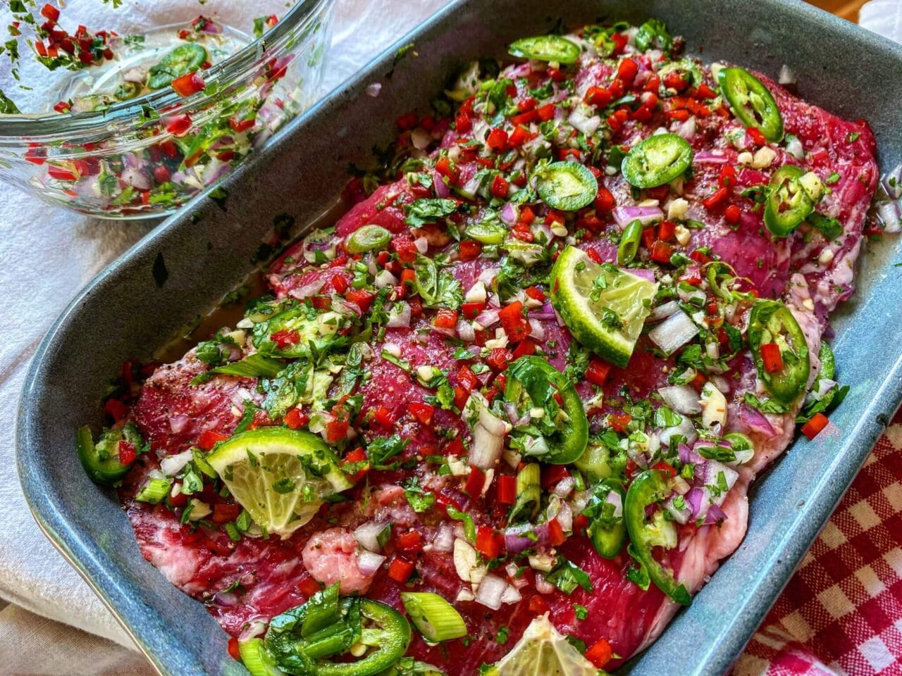 A marinated piece of raw meat in a baking dish, topped with chopped jalapeños, red peppers, onions, cilantro, and lime wedges.
