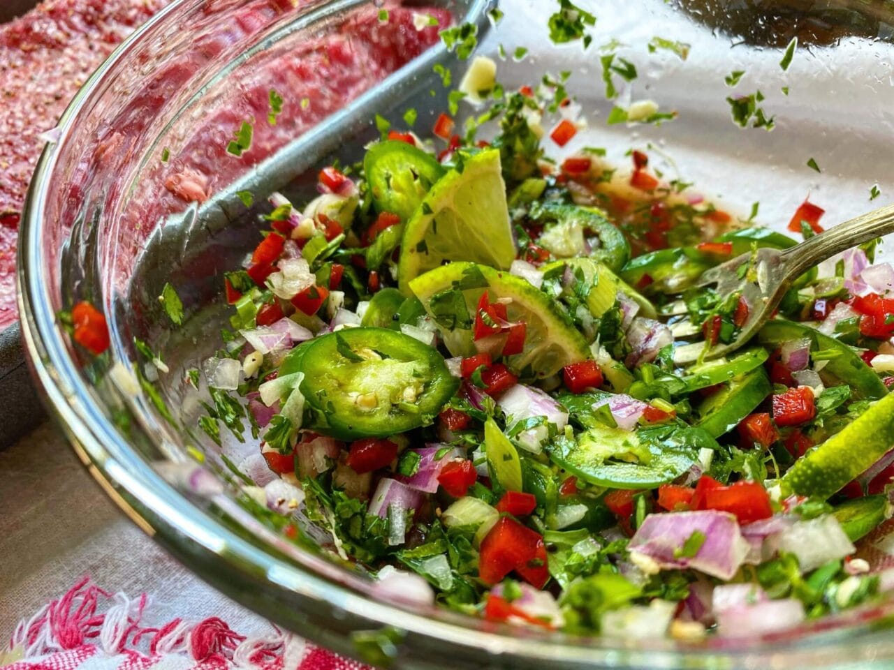 A glass bowl of chopped vegetables with jalapeños, red peppers, onions, cilantro, and lime wedges.