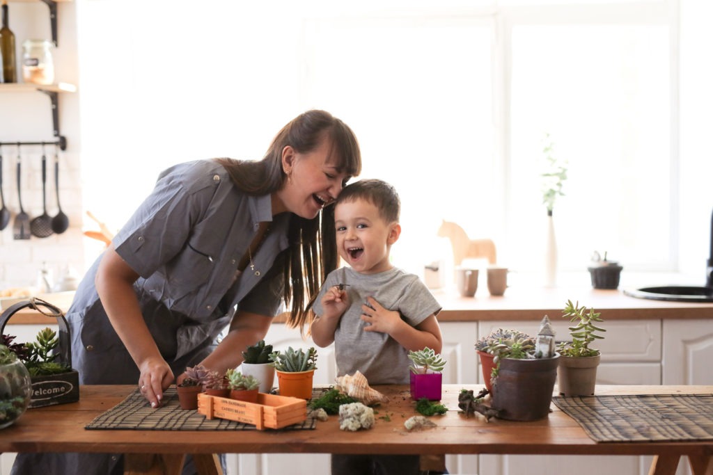 Cute child boy helps his mother to care for home plants. Mom and her son engaged in gardening near window at home. 