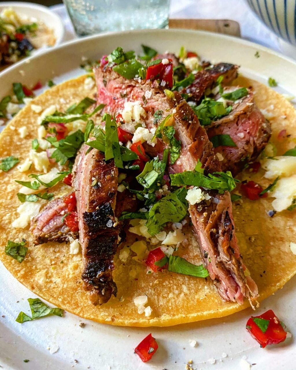 A taco with grilled meat strips, chopped cilantro, red peppers, and crumbled cheese on a corn tortilla.